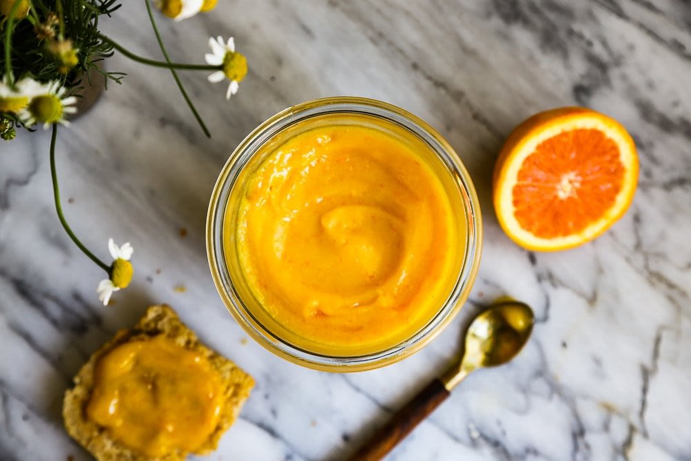 An orange curd in a glass jar on a marble table surrounded by an orange, spoon and flowers.