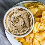 A bowl of vegan sour cream and onion dip surrounded by crispy potato chips.