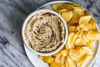 A bowl of vegan sour cream and onion dip surrounded by crispy potato chips.