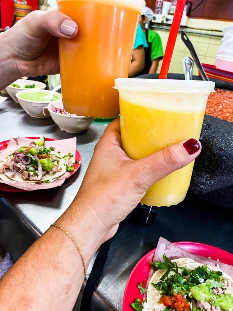 Two brightly-colored fruit juices held in the air by two hands over a restaurant counter with tacos on it.